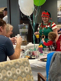 A group of people sitting at a table, a little kid is looking a girl wearing an elf costume. Red, green, and white balloons on the table.