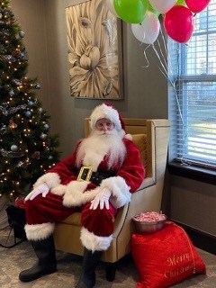 A man wearing a santa costume is sitting in a chair next to a christmas tree and a big red bag full of candy canes. Red, green, and white balloons on the right.