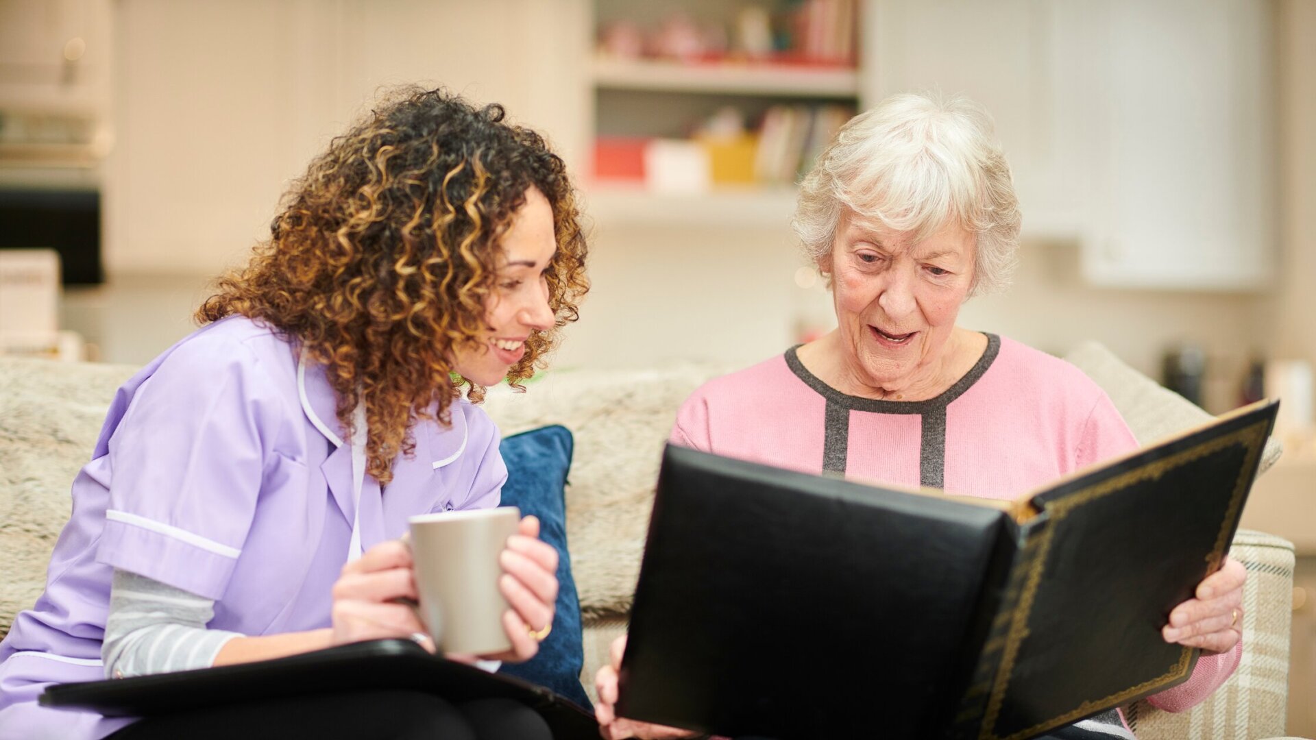 A woman in purple scrubs onlooks an older person in white reading a book