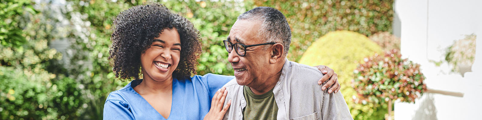 A senior man walking outdoors with his female nursing assistant as she puts her arms on his shoulders and smiles