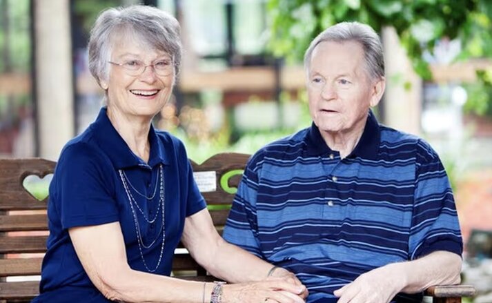 Husband and wife sit together on a park bench holding hands and smiling at Traditions at Reagan Park in Avon, IN.