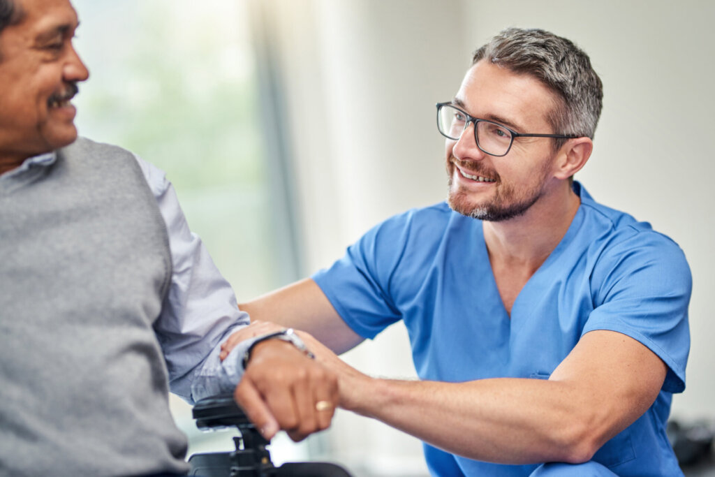 Male nurse sitting next to a male memory care resident, laughing and conversing together at Traditions at Regan Park in Avon, IN.
