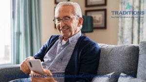 An elderly man is wearing glasses and blue jacket is sitting on a gray couch looking down at his phone.