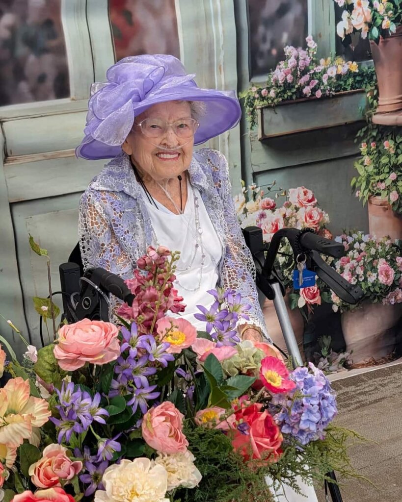 A beautifully dressed resident smiles amid a colorful floral arrangement at a garden tea party event at Traditions at Reagan Park.