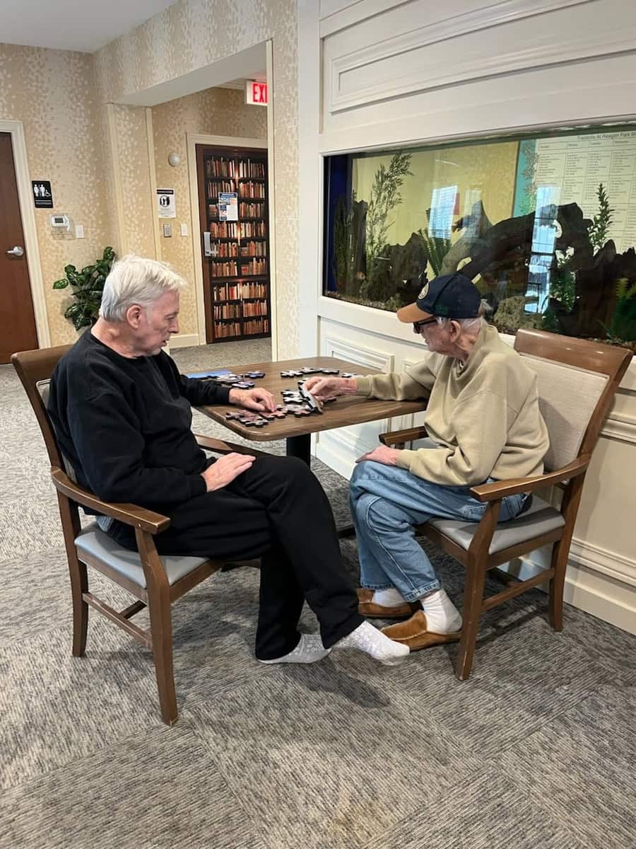 Two residents work together on a jigsaw puzzle in the common area at Traditions at Reagan Park, with the community aquarium visible in the background.